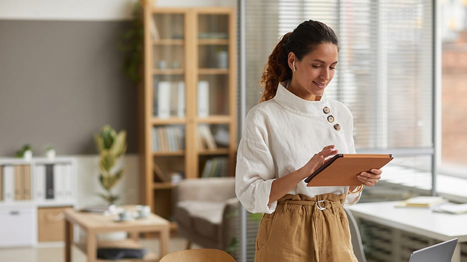 Portrait of elegant businesswoman using digital tablet with wireless earphones while standing by desk in office or at home, copy space