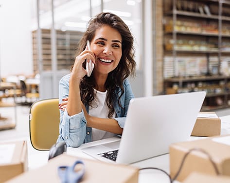 Happy young businesswoman speaking on the phone while working in a warehouse. Online store owner making plans for product shipping. Creative female entrepreneur running an e-commerce small business.